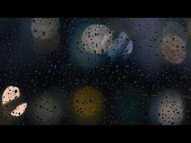 A night photo of a window glass with raindrops scattered on the surface with very blurred city lights behind it.