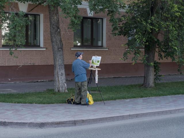 A photo of somewhat aged man painting a picture. He's wearing a blue t-shirt with white horizontal stripes, khaki camouflaged pants and a summer baseball cap. In front of him there is a tripod with a box with paints and brushes attached to it. Above the box there is an unfinished picture. In the picture we can see trees and some old buildings between them. In the background of the photo there are a row of trees and a ground floor of a relatively old building in quite good condition with new windows and white vertical blinds behind them.