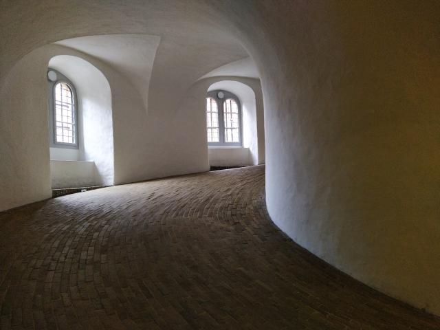 Curved path up the inside of a tower. The walls and ceiling are white render. The floor is individual narrow paving blocks maybe fifty of them across the width of the path all pointing with the long edge along the path. The path curves around to the right and out of sight. In the left hand wall are two arched recesses each with a double arched window to the outside.