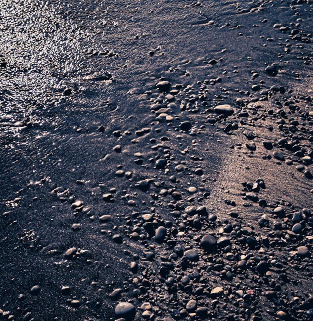 A beach. The water is just coming in on the left of the frame. The right of the frame is small rocks in the sand. Water glints off of the water and wet sand. 