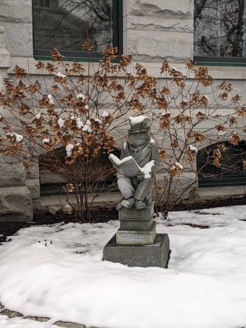 A grey stone statue of a sitting mouse wearing a suit and top hat and holding an open book.
Around the statue on the ground: fluffy white snow. Snow also sits on the top rim of the book, the mouse's arms and shoulders, and on the top hat. 
Behind the statue: spindly trees with brown/orange leaves littered with snow.
All in front of a building with reflective windows and uneven tan stone.