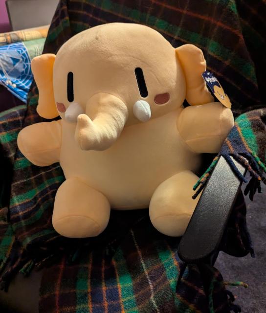 Pale beige plushie mastodon sitting on a chair. It is a beautifully peachy soft, chunky bodied plush toy. It has little blush cheek patches, & white tusks. It sits on my chair, which is draped with a Fraser tartan blanket.