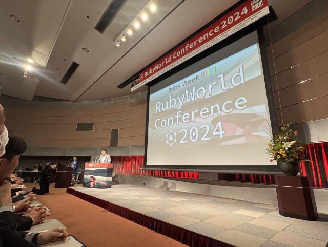 Matz is standing to the left of a stage with a slide on the screen that reads "RubyWorld Conference 2024" 