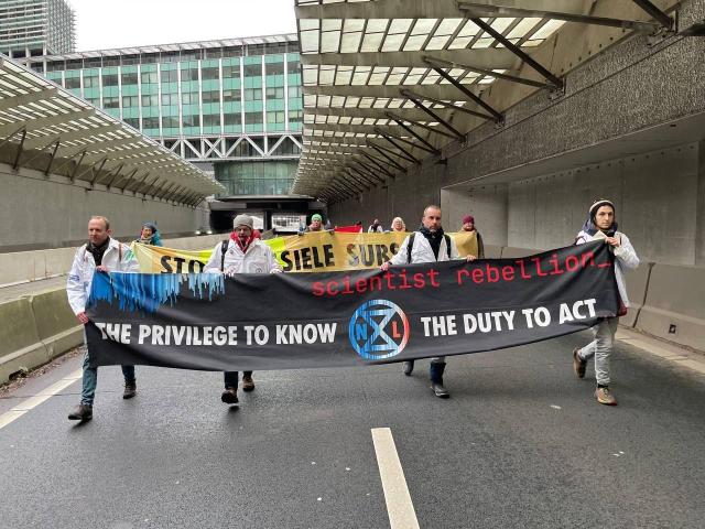 Four activists from Scientist Rebellion walk on the A12 highway in The Hague. They are wearing white lab coats, and hold a banner saying 'The privilege to know, the duty to act'. In the background, more banners and demonstrators are visible.