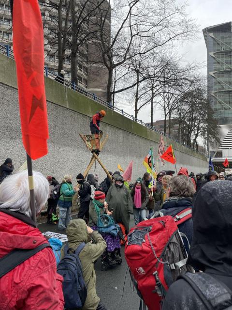 Demonstrators are sitting and standing on the A12 highway in The Hague. Some of them are holding flags. One demonstrator wearing a helmet is standing on a crow's nest made from bamboo poles, displaying a flag with Extinction Rebellion's logo on it.