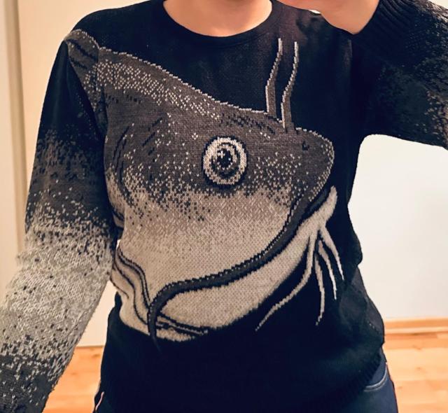 Photo of me wearing a black sweater that has a grayscale knitting pattern in the shape of a catfish head. It resembles pixel art. 