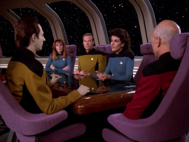A TNG scene. We're in the Enterprise conference room, where Picard is holding a meeting with Data, Troi, and Dr Crusher. Barclay is also there, for unknown reasons. Maybe he wandered into the meeting and just sat down, and everyone was too polite to mention it. That happened to me once. I was visiting an office location I didn't normally go to and I wasn't quite sure which conference room I was supposed to be in. I walked into one and sat down and it took me five minutes to realize I was in the wrong meeting. From the perspective of the people in that room, halfway through that meeting, a stranger walked in, sat next to the boss, took notes for five minutes, then walked out without saying a word.