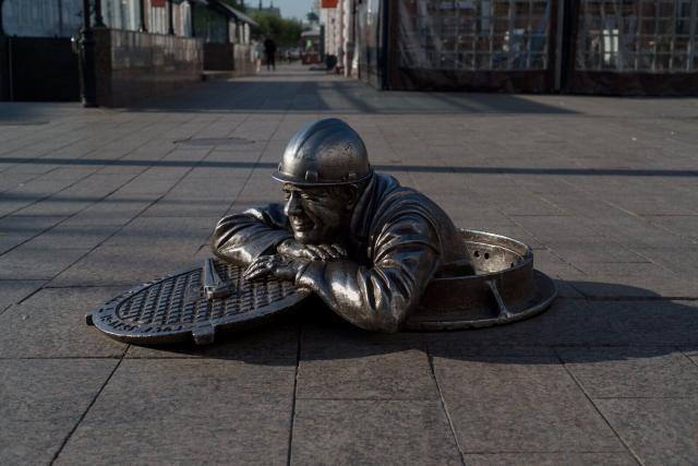 A photo of a pedestrian street with a sculpture n the ground. The sculpture is a portrait of a plumber, a middle-aged man in a working robe and a helmet leaning out of the manhole. Only his head, shoulders and hands are outside. He leans calmly on the side of the manhole with the lid of the manhole under his arms, his head on his hands. On the lid there is a plumber wrench. He looks forward with a calm smile.