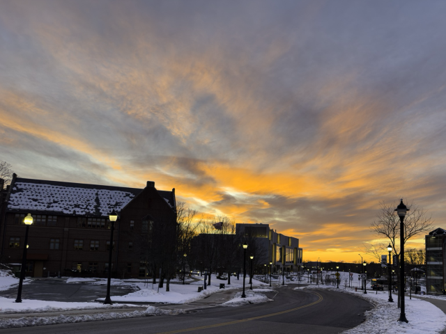 Orange clouds over a darkened street with streetlamps along the side and two large buildings. 