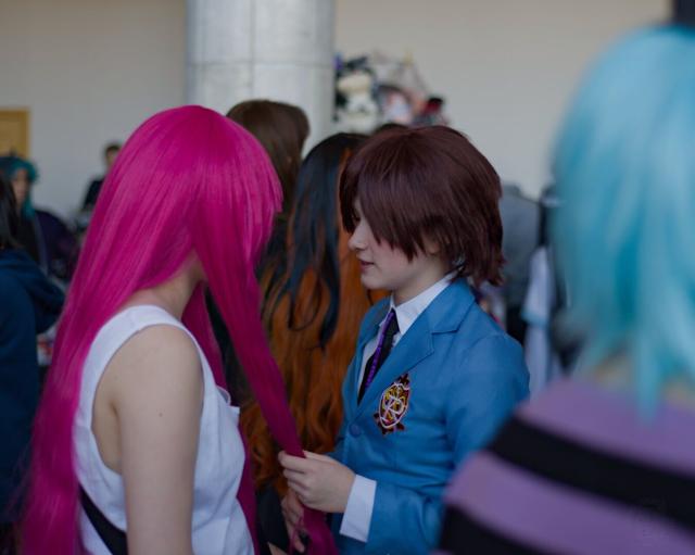 A photo of 2 persons at cosplay event. A girl on the left is wearing white blouse without sleeves and a wig with long pink hair. She photographed from her right and back, and her face is not visible. A girl on the right is wearing a white shirt, black tie and bright-blue jacket with red and gold embroidered emblem with white letters “O” and “R” on it. She looks to the first girl and softly holds in her hands one strand of the pink wig. They both stay in the middle of a crowd of other visitors.