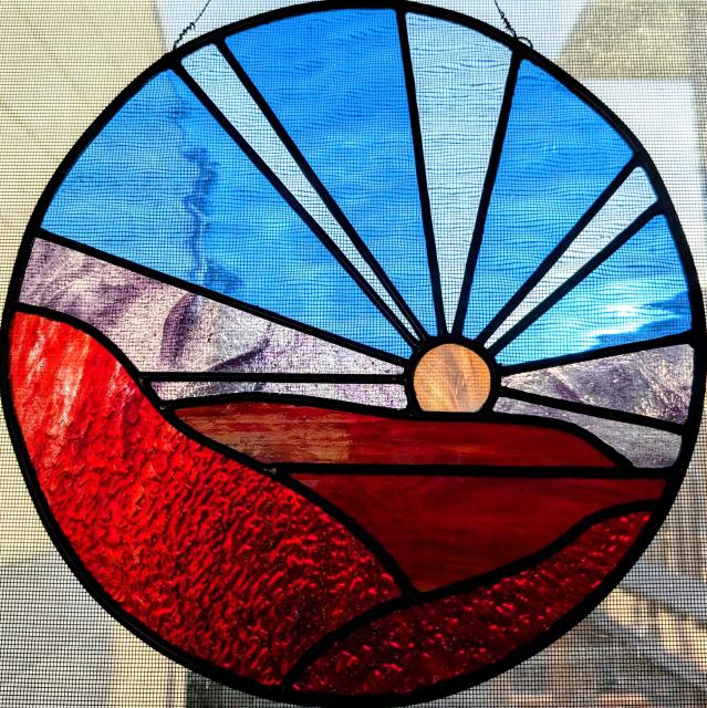 Photograph of a round stained glass window hanging. The scene is a Martian landscape: red rocky foreground and darker red distant mountains with a white sun on the horizon (it looks more yellow in this photo because it was taken during golden hour on a cloudy day and the actual sun was low in the horizon; my camera angled to catch the muted rays of the sun through the stained glass). The stained glass sky is blue (with a dusky purple near the horizon), evoking a blue Martian sunset. CC BY-SA Kate Zimmerman.