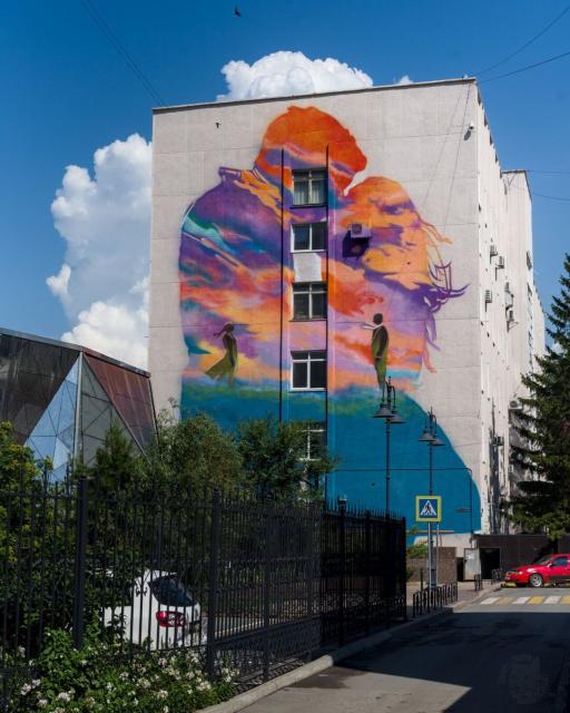 A photo of a 5-storey building with a mural on its wall. Mural background is a silhouette of a man and a woman kissing each other filled with an abstract colorful spots resembling a landscape with green grass, blue mountains near the horizon and wild purple and orange sky during a sunset. In the foreground there are silhouettes of a man and a woman standing far from each other and looking to each other.