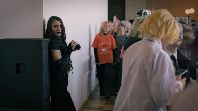 A photo of a woman with long wavy black hair dressed in all black leaning against a white wall and looking straight to the camera. There are other people in the frame, they are mostly teenagers in cosplay costumes, they are out of focus. There is a visible distance between the woman in black and the other people.