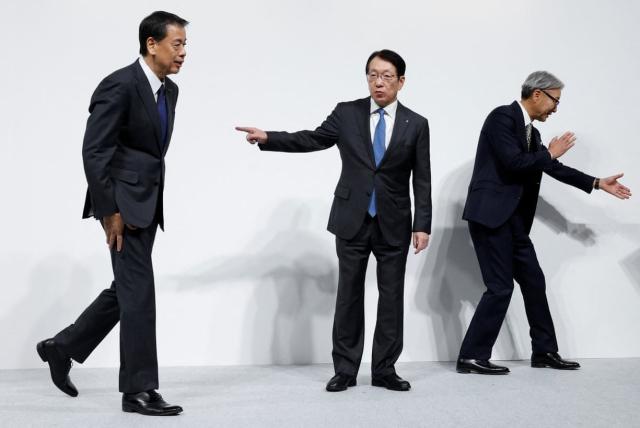 Three Japanese men in business suits standing against a stark white background. They each have a distinctive pose: the one on the left is walking with hands by his side, the one in the middle is turned to the one on the left and is pointing at him with a single outstretched hand, and the one on the right is turned away from them both, gesturing at something out of shot with both hands.