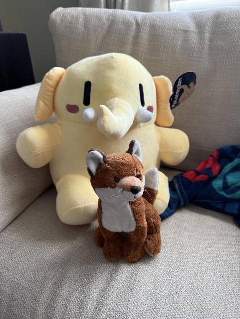 Yellow Mastodon plush mascot sitting on a white couch with a smaller fox plush in front and a CBS blanket to the side.