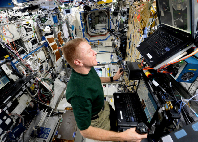 screenshot of astronaut on ISS at a computer workstation. Behind them is a whole mess of tech and wires.
