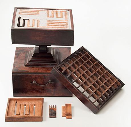Image of an old wooden incense clock, a box with a top section containing sand with incense pattern drawn onto it that will slowly burn over a fixed period of time, a lid with square holes goes on top