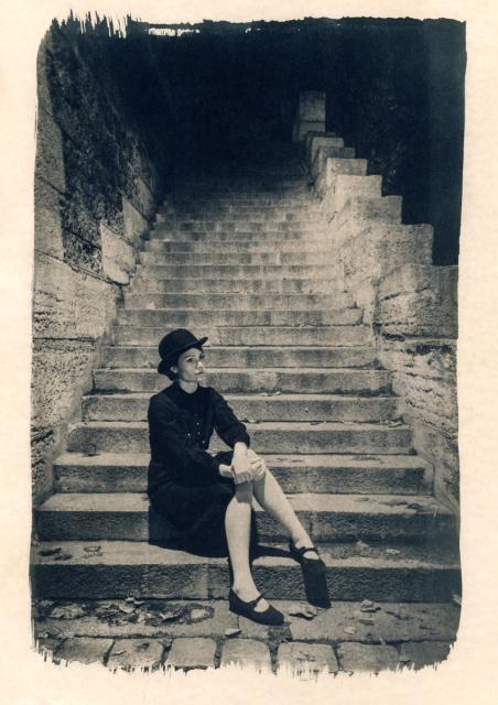 A woman dressed in a 1930s style and wearing a bowler hat is sitting at the bottom of a long flight of stone stairs in central Paris. The image looks painted from the cyanotype and sepia from the coffee toning. 