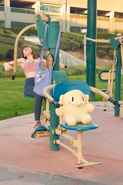 Photo of Lety and the Creature Mastodon stuffed toy on some outdoor fitness equipment near sunset. From top to bottom, Lety is seen wearing a mauve sports bra, gray leggings, and light pink boots. She's sitting in the back half of a piece of equipment labeled “2-Person Back & Arms Combo”, her feet on footrests in front of her as she orients her elbows parallel to her shoulders to pull back on giant metal levers that lift her seat. The Creature is seated in the front half of the equipment, which is very similar but with levers intended to be pushed down for lift instead of pulled back. In the background behind them are more fitness equipment, then a grassy area followed by some uphill shrubbery and a multi-level parking garage.