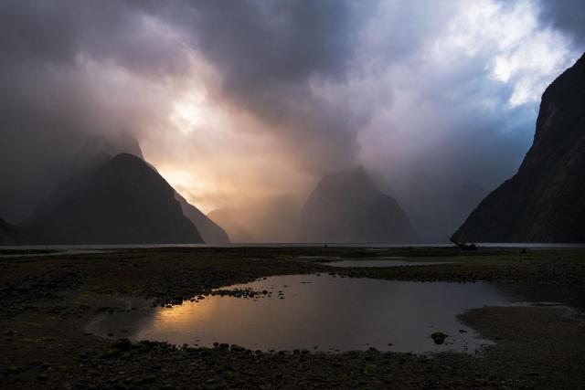 a stormy sunset in Milford Sound in NZ with dark menacing clouds and some light shining from behind the mountains