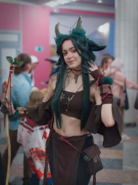 A photo of a young woman wearing brown top and sort of black pants with brown apron from waist down. She has long green hair. small horns and green ears similar to cow or deer ones. In her right hand she holds sort of staff made from smooth yellowish wooden stick without a bark with several green leaves on top. Her left hand is near her face. She looks to the camera and smiles.