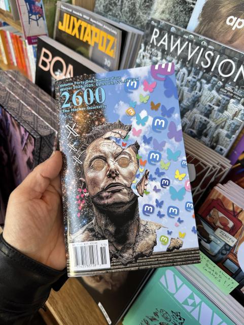 A person holding a magazine titled "2600 The Hacker Quarterly," featuring a surreal cover with a cracked sculpture of a face and colorful butterflies against a cosmic background. Other magazines are visible on the shelf in the background.