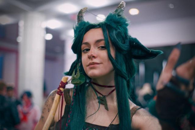 A head and shoulders portrait of a young woman wearing brown top and red choker with a pendant in the form of the first rune of futhark (fé/fehu/feoh). She has long green hair. small horns and green ears similar to cow or deer ones. In her right side some sort of staff is visible, it's made from smooth yellowish wooden stick without a bark with several green leaves on top. Her left hand is stretched towards the camera and is out of focus. Since she photographed a bit from below, she looks to the camera with her eyes softly closed with an expression similar to smug.