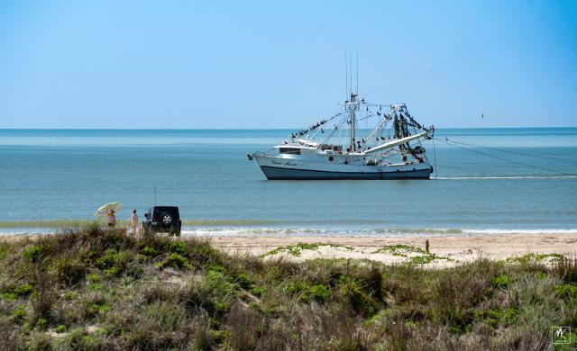 Color photo of a Gulf of Mexico shrimp boat trawling for Gulf of Mexico shrimp just offshore of Matagorda Beach located on Texas’s Gulf of Mexico coast. There are two people on the beach next to a Jeep setting up an umbrella. Gulf of Mexico in the background.  