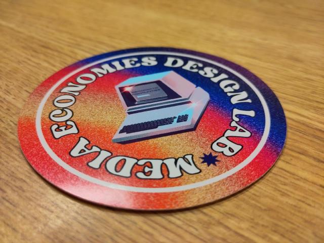 Colorful sticker that says Media Economies Design Lab in a circle around an old computer that says "democratic communities" on the screen