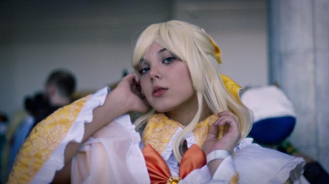 A head and shoulders portrait of a young woman wearing a yellow dress with long and wide sleeves and with white frilly cuffs. On her chest a big red bow is visible. Her yellow-white hair is shoulder-long, and decorated with yellow bow on the back of her head. Her right hand almost touches her right ear, and her left hand is half-way to her face. She looks to the camera a bit to-down with her mouth slightly opened. Her eyes are steel-gray with bluish tint.