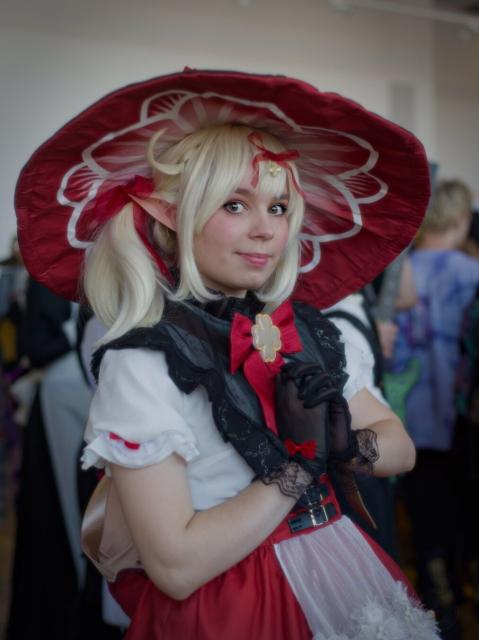 A waist-height photo of a young woman with white-yellow hair tied in pigtails. She wears a red witch hat with white linings, a white blouse with buttons, a red ruffled dress with girdles, white knee-high socks along with 3 small red bows on the sides, black gloves, and Mary Jane shoes which have small gold flowers with four petals attached to them. Ribbons and clover decorations are embellished throughout. She also wears a large leather bag. Her hands are closed at a chest level.