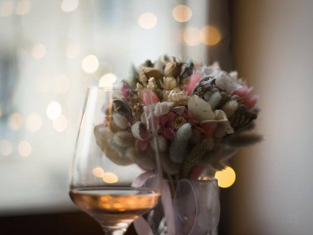 A photo of a bouquet made of dry flowers in pastel tones. In the foreground there is a blurred tall and wide wineglass with a little of pink wine in it. Background is mostly very blurred little sparks from LED garland hanged on a window.