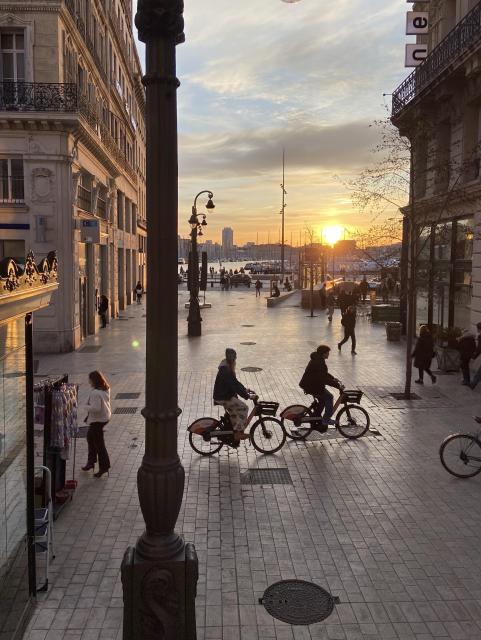 The sun is setting between two buildings. Two people are bicycling past and a few other people are standing or walking 