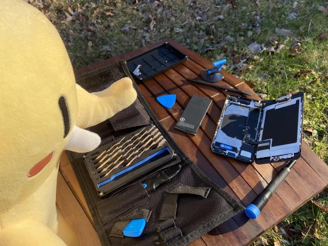 Mastodon plush hovering above a small wooden table that's outside on the grass. On the table is an iFixit toolkit and iPhone SE (2022) with its battery removed. The tools and phone components are strewn across the table mid-repair.