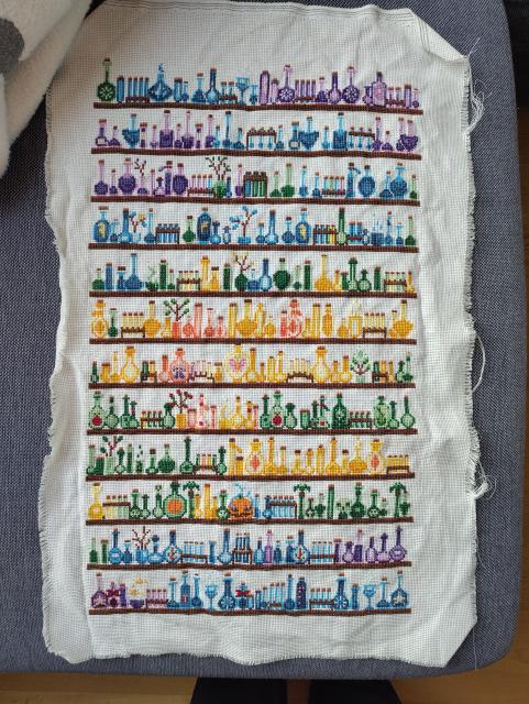 Huge cross stitch piece on white fabric: twelve shelves of little portions in shades of blue, purple, green, yellow, and orange. Some potions are seasonally decorated with plants, stars, or skulls. The fabric is wrinkled and a little frayed.