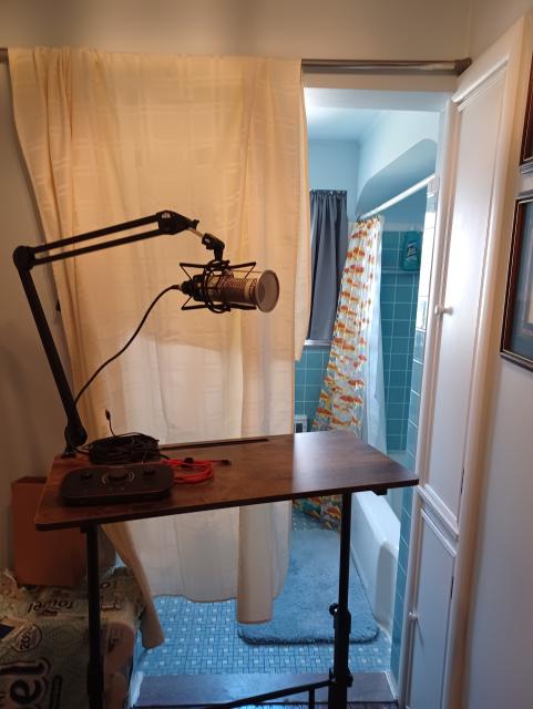 my portable standing desk with mic mounted on arm. behind it, a hanging tablecloth partially obscuring the rest of the bathroom.
