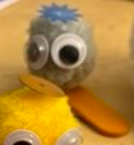 Smaller of the pompon-based creatures with splayed half-popsicle-stick feet. It features: googly eyes that are almost too large for its body; a flat, spiky blue hat; and orange feet.