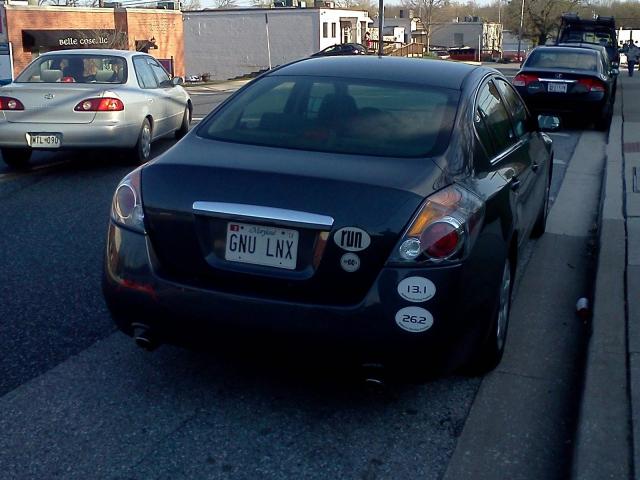 A black car is parked on the street. The license plate reads GNU LNX.  GNU Linux!  Penguins everywhere!  Well, no.  No penguins.