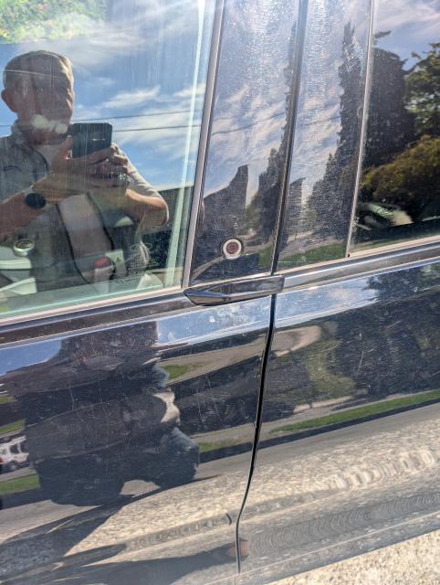 Photo of me in front of a Ford Mustage Mach E. It shows the "door handle" which is just an enameled metal flange that doesn't move. You open the door by pushing a button, which pops open the door.