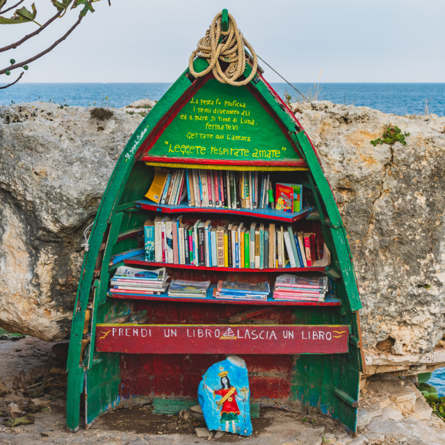 A photo of a public bookcase made from the front half of a small boat, standing upright against a rocky background with the sea in the distance. The boat is filled with various books, and there’s a sign in Italian that reads, “Prendi un libro lascia un libro,” which means “Take a book leave a book.”   