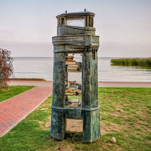 A photo of a public bookcase resembling a lighthouse with books inside and the word “Biblioteka,” which means library in Lithuanian, at the top. It’s placed on a grassy area next to a red brick pathway leading towards the sea.   
