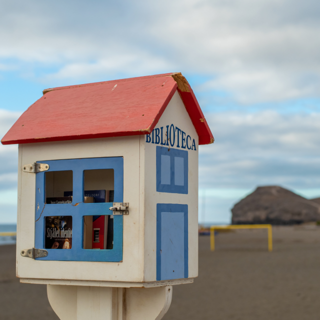 A photo of a little bookcase labelled "Biblioteca," which means “library” in Spanish. It resembles a small house with a red roof and blue trim, and it’s filled with books. In the background is the beach and the ocean.   