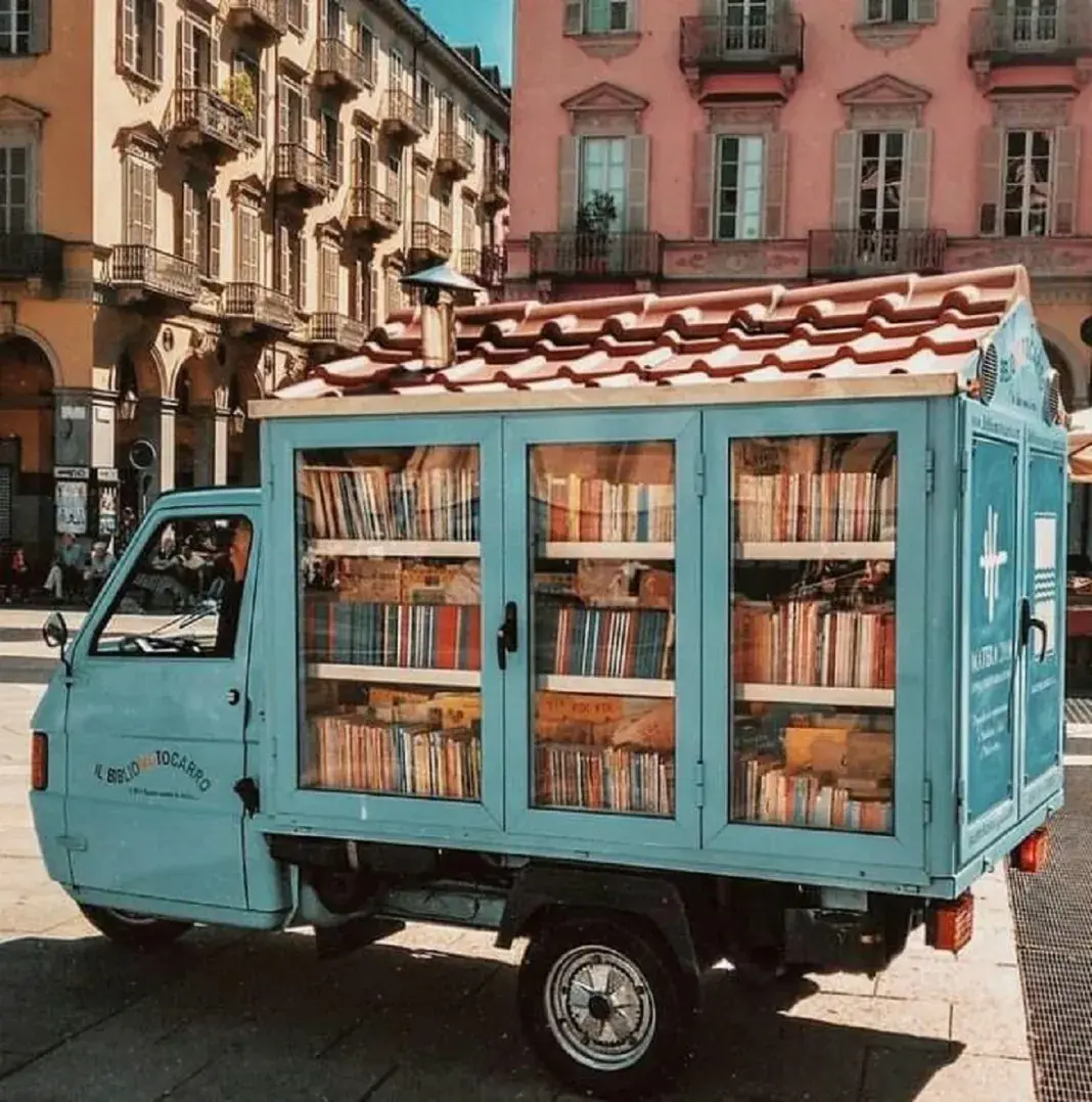 A charming light blue three-wheeled van-motorbike transformed into a mobile library, known as the "Bibliomoto," parked in a sunny Italian square. The vehicle's rear is fitted with glass-paneled wooden shelves packed with colorful books, topped with a red-tiled roof resembling a miniature house. The scene is set against elegant historic buildings in the Basilicata region of southern Italy, where the Bibliomoto brings literature to remote villages.