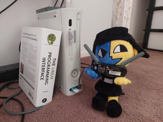 a phat Xbox 360. a thick book called "the Linux programming interface" leans against it. a plushie of the character ENA holds the wifi module, which is connected to the 360 via USB. the entire thing is on a carpet.
