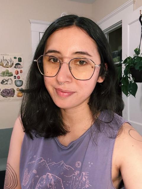 person with olive skin and dark shoulder length hair wearing glasses and a purple tank top, smiling at camera. in the background is a hanging plant and a poster of gemstones