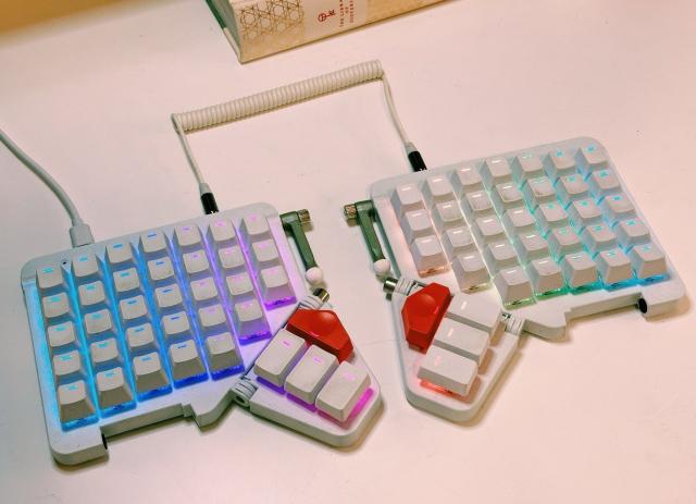 a white split ergonomic keyboard with rainbow backlighting, unlabeled keys, and red thumb buttons, resting on a white table