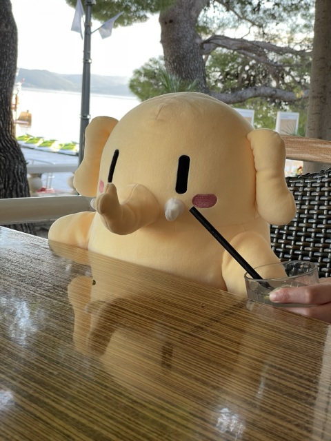 A stuffed Mastodon toy sitting in front of a cafe table outdoors, with sea behind him. A glass of lemonade is in front of him.
