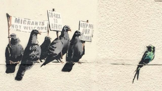 5 pigeons with signs "Migrants not welcome", "Go back to Africa", "Keep off our worms" protesting against a swallow.