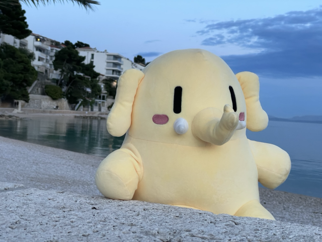 A stuffed Mastodon toy is in front of a a Mediterranean beach during dusk.