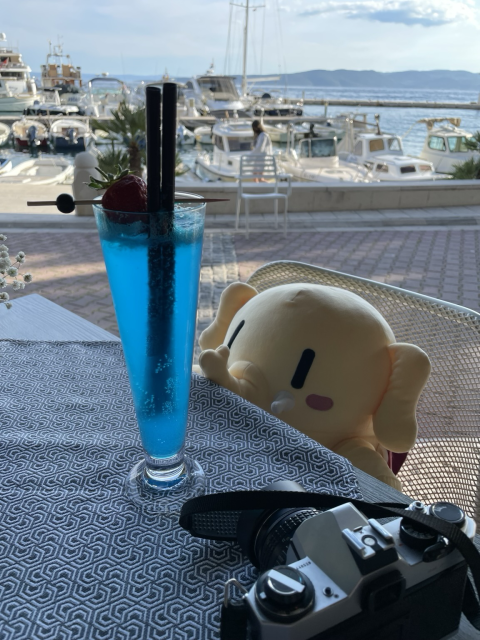 A stuffed Mastodon toy peeking at a blue cocktail from behind the cafe table, with the sea in the background.